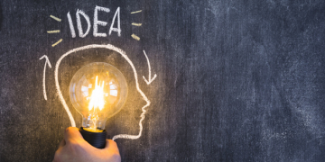 hand holding illuminated light bulb with outline face drawn on blackboard with idea text 23 2147874059