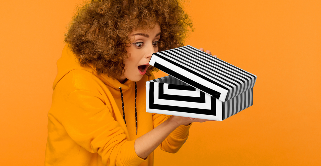 Unboxing extremos: una nueva estrategia de marcas e influencers para vender en redes sociales 1 Portrait of funny, surprised, curious curly-haired woman in hoodie looking into gift box, unwrapping present and peeking inside with interest, open mouth in amazement. indoor studio shot isolated