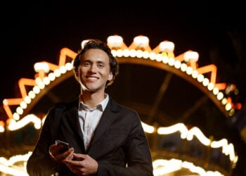 Portrait of young successful businessman in suit looking at camera, smiling, holding phone over night amusement park.