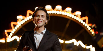 Marketing de Entretenimiento: ¿Cómo conectar marcas y emociones en la era digital? 2 Portrait of young successful businessman in suit looking at camera, smiling, holding phone over night amusement park.