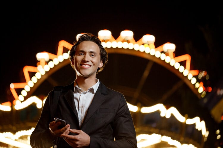 Portrait of young successful businessman in suit looking at camera, smiling, holding phone over night amusement park.