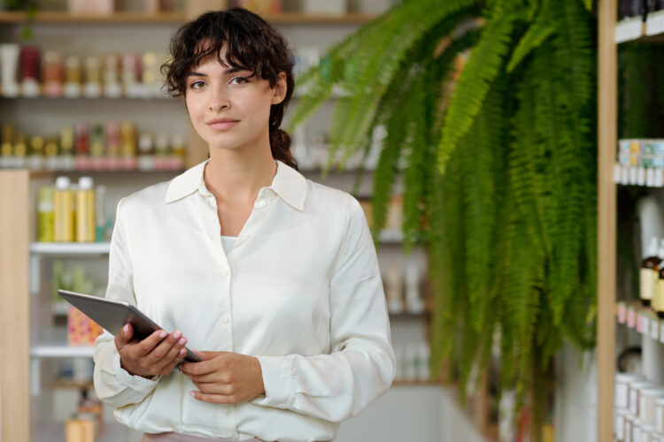 Young woman in smart casualwear standing in cosmetic shop or supermarket against display and looking at camera